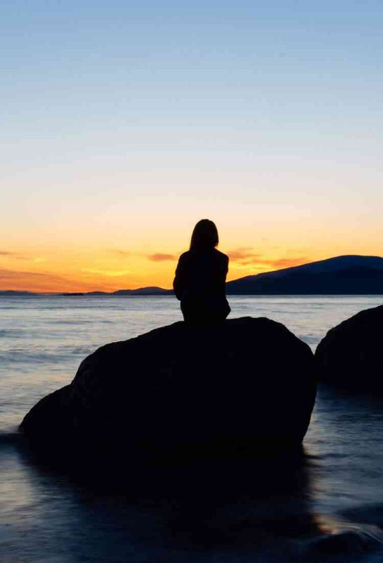 Silhouette of a woman meditating on ocean rocks at sunset with glowing sky.