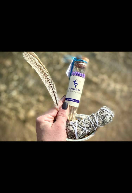 Hand holding Nymph Ki intention matches, feather, and sage bundle against a natural outdoor backdrop.