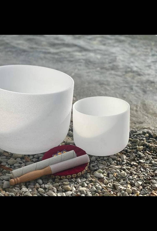Two white crystal singing bowls with mallets resting on a bed of beach pebbles by gentle waves