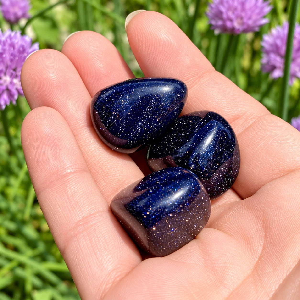 The shimmering Blue Goldstones in an open palm over blooming chives with purple flowers and green stalks.