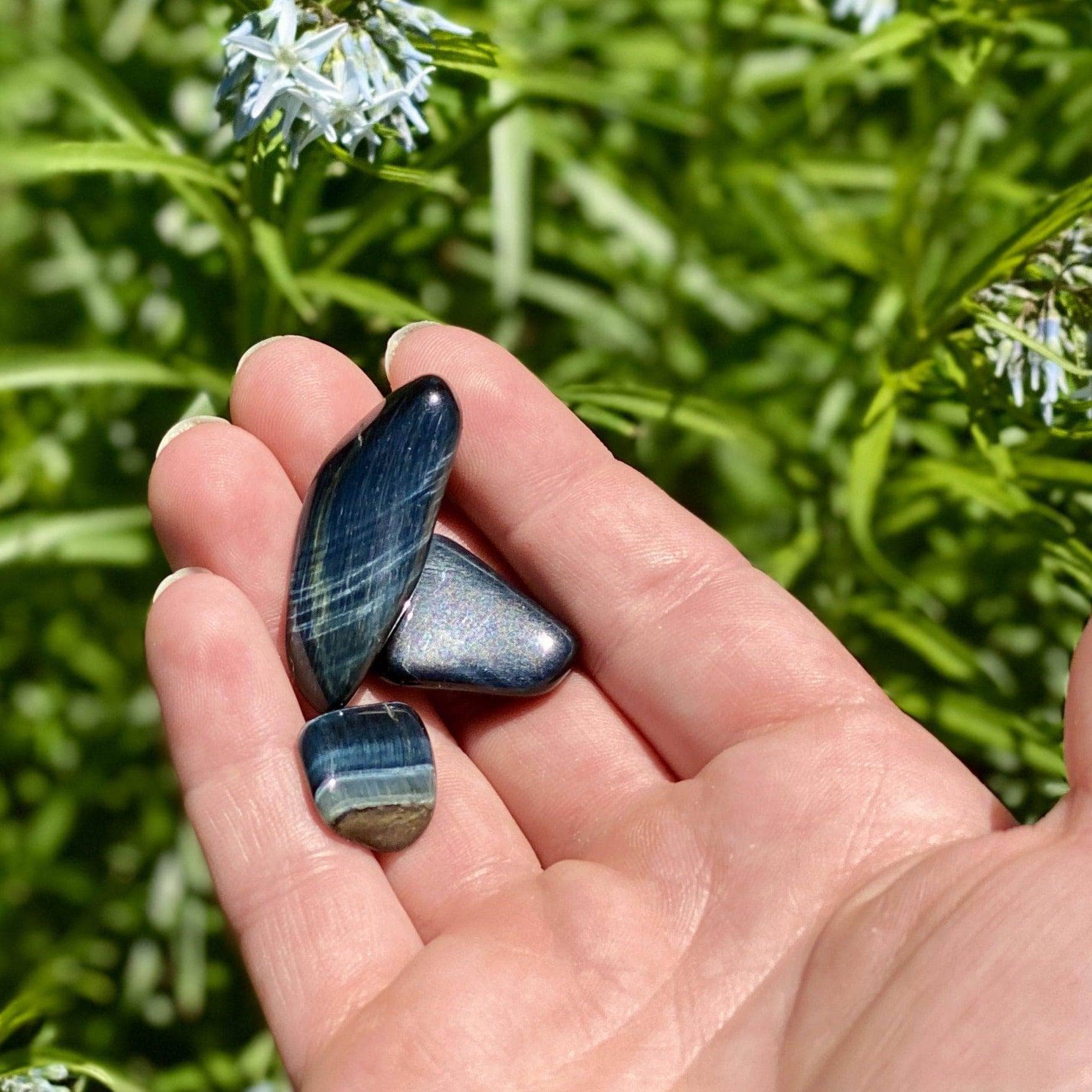 Three polished Blue Tiger’s Eye stones resting in an open palm with soft green grass in the background.
