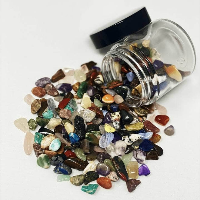 Mixed chakra healing stones spilling from a glass jar in a vibrant rainbow of crystals on white background.
