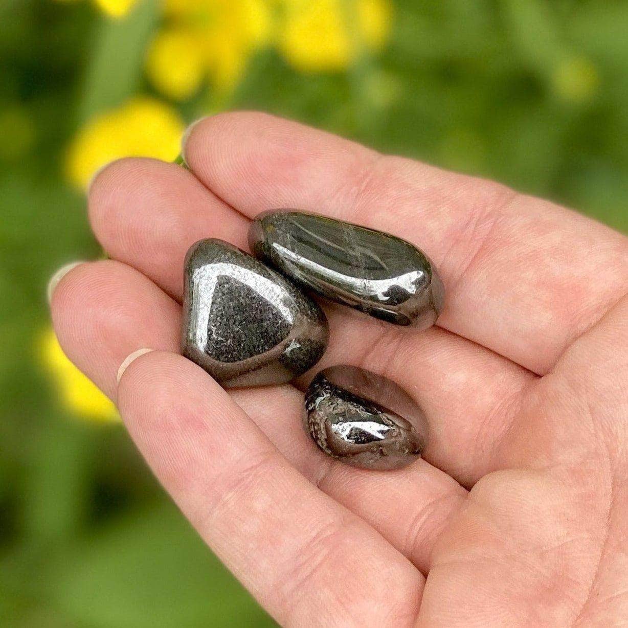 Three polished hematite stones resting on an open palm with bright yellow flowers in the background.
