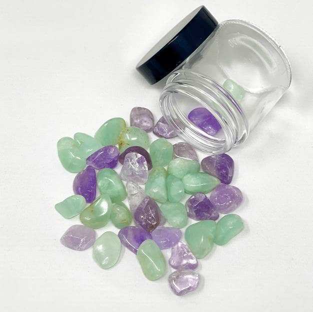 Amethyst and Prehnite stones spilled from a glass jar in a soft green and purple mix on white background.
