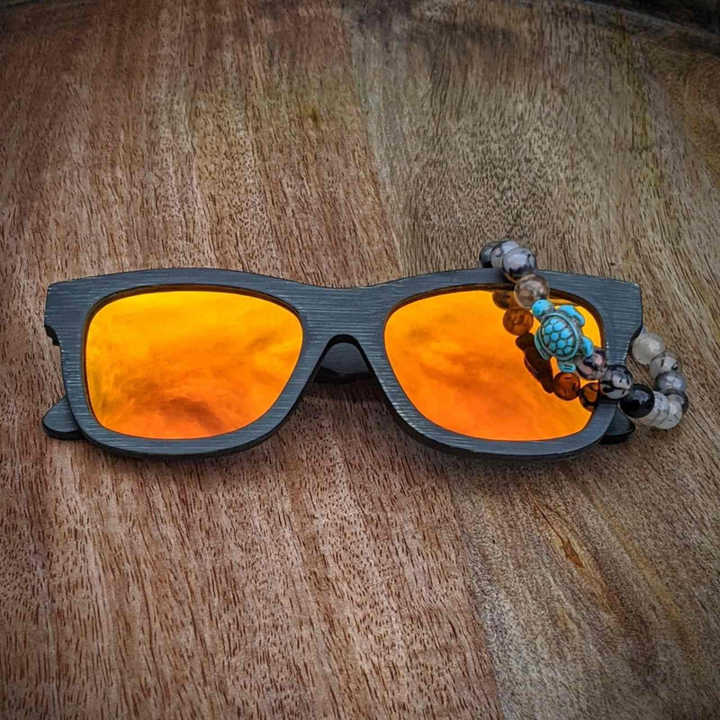Black bamboo sunglasses with orange mirrored lenses and a qurtz  bracelet featuring a turquoise turtle charm resting on wood.
