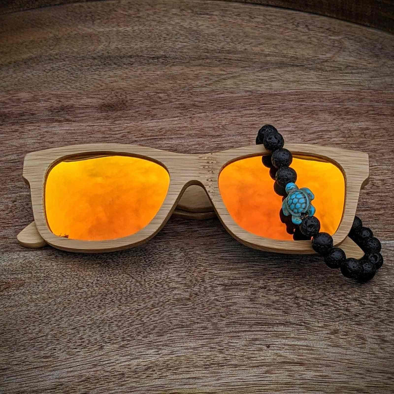 Natural bamboo sunglasses with orange mirrored lenses and a lava stone beaded bracelet featuring a turquoise turtle charm resting on wood.
