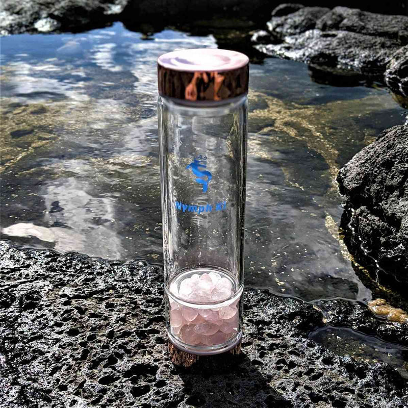 Nymph Ki water bottle filled with rose quartz gemstones reflecting the sky in a tidepool.
