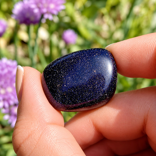 Close up of one Blue Goldstone crystals sparkling in fingers above blooming purple chive flowers.