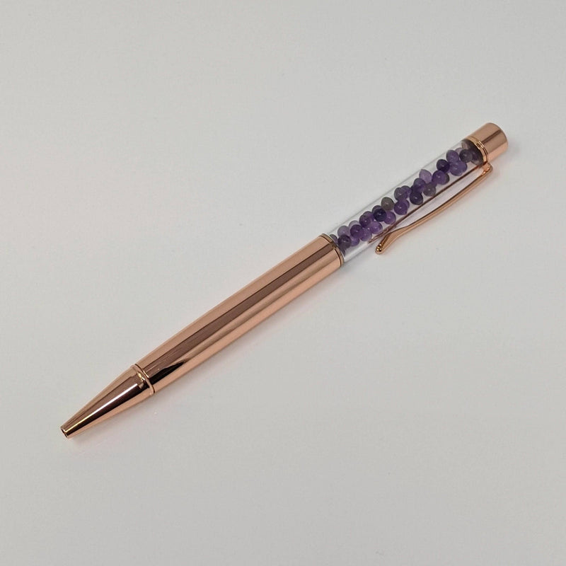Full-length flat lay of rose gold pen filled with amethyst crystals, shown on white background.
