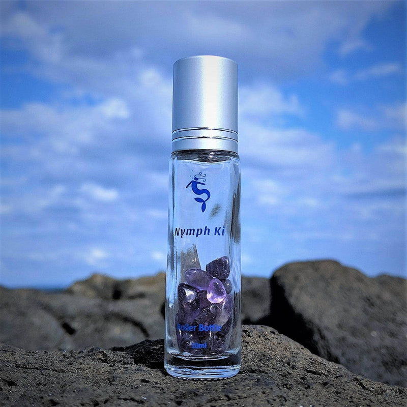Close up of Crystal roller bottle displayed on volcanic rock with vibrant blue sky and clouds behind it.
