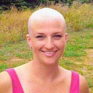 Smiling bald woman in a pink top standing outdoors in front of grass and trees.
