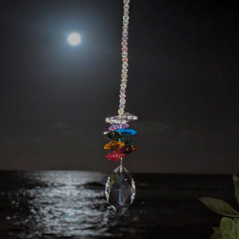 Chakra crystal suncatcher glowing under the full moon above ocean waves, with a prism pendant and rainbow beads.
