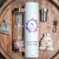 Nymph Ki Hope Gift Set with rose quartz crystal water bottle, travel tube, stainless steel infuser, chakra sun catcher, and two crystal bracelet pouches on a wooden tray.
