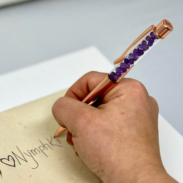Hand using rose gold pen with Amethyst crystals to write “Manifest” on seeded paper.