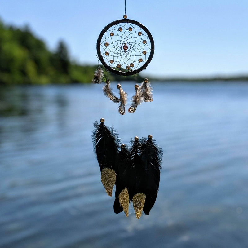 Black dreamcatcher with gold-tipped feathers and a beaded center hanging above a calm lake on a sunny day.
