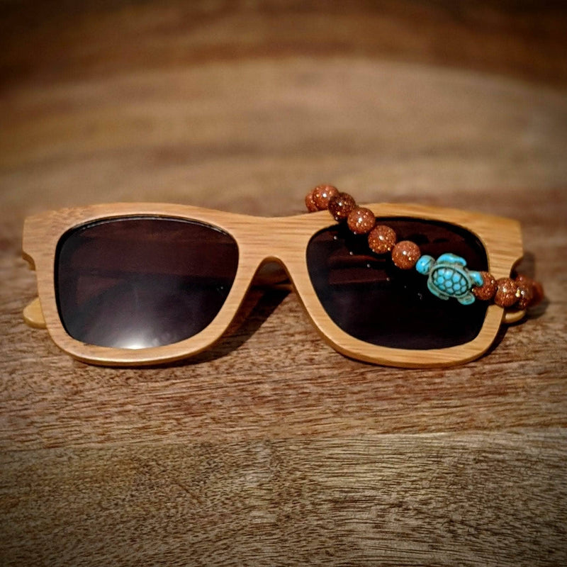 Black lens bamboo sunglasses paired with a gold sandstone turtle bracelet, displayed on a warm wood surface.
