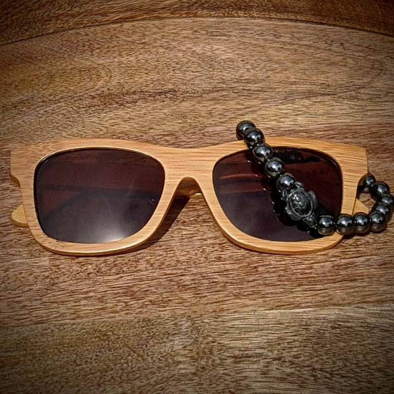 Black lens bamboo sunglasses paired with a polished hematite turtle bracelet, displayed on a smooth wooden surface.
