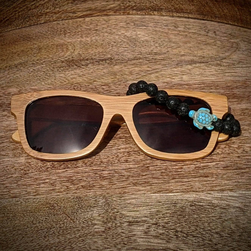 Black lens bamboo sunglasses paired with a lava rock turtle bracelet, displayed on a warm wooden background.
