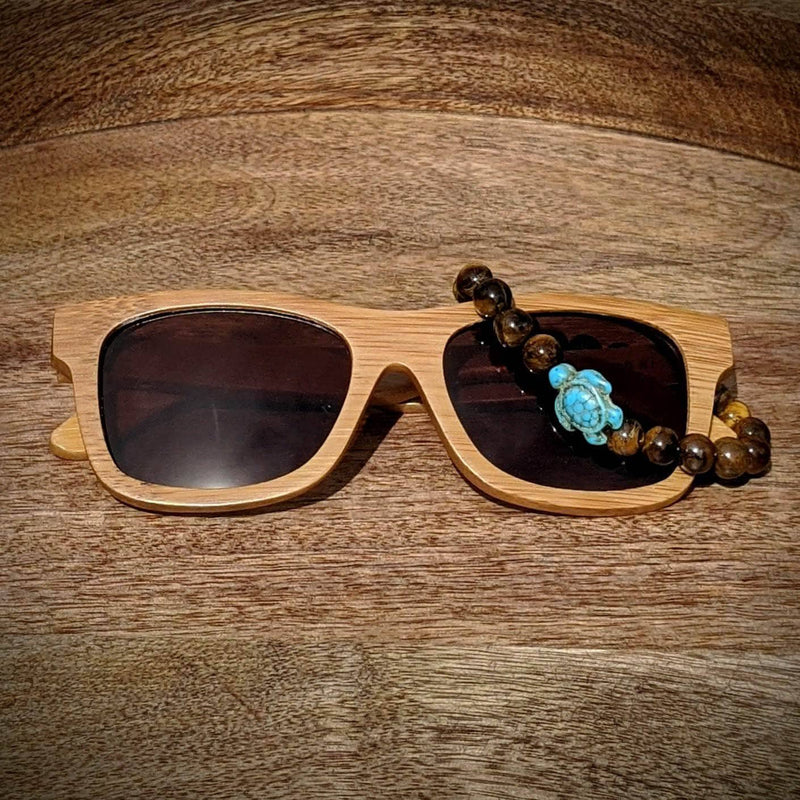 Black lens bamboo sunglasses paired with a tiger’s eye gemstone turtle bracelet, displayed on a warm wooden surface.
