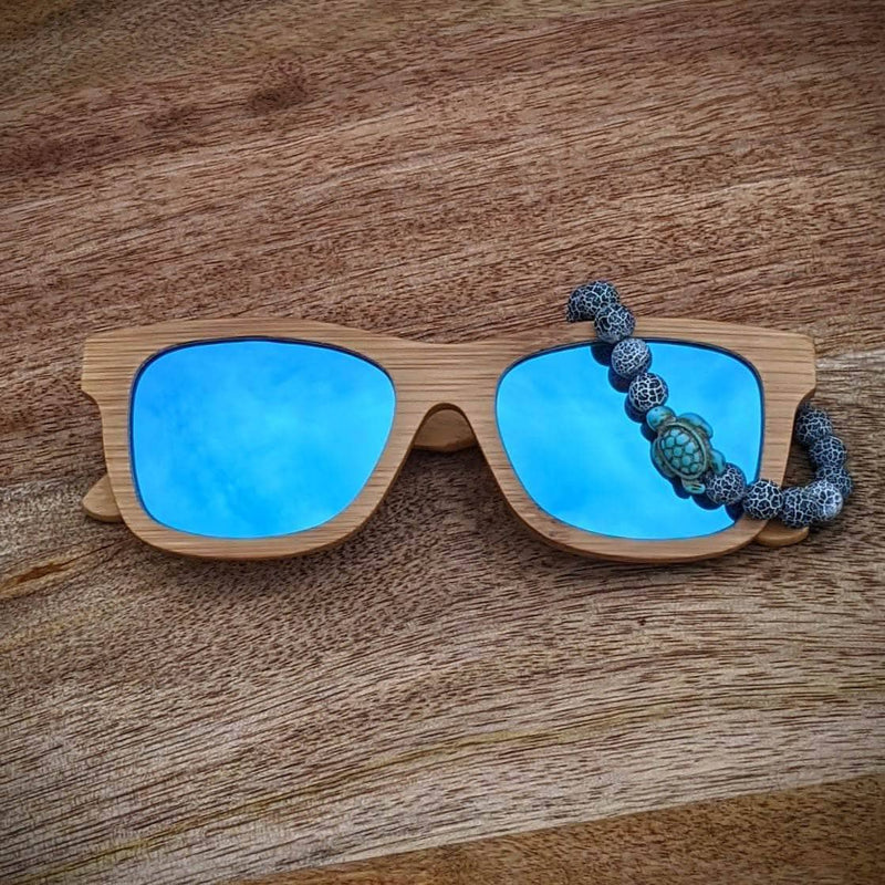 Blue mirror lens bamboo sunglasses paired with a gray lava rock and turquoise turtle bracelet, displayed on a round wooden surface.
