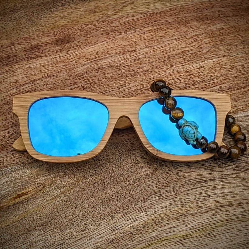 Blue mirror lens bamboo sunglasses paired with a tiger’s eye gemstone turtle bracelet, displayed on a rustic wooden surface.
