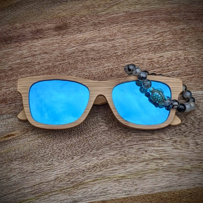 Blue mirror lens bamboo sunglasses paired with a Tourmalinated Quartz turtle bracelet, displayed on a natural wood surface.
