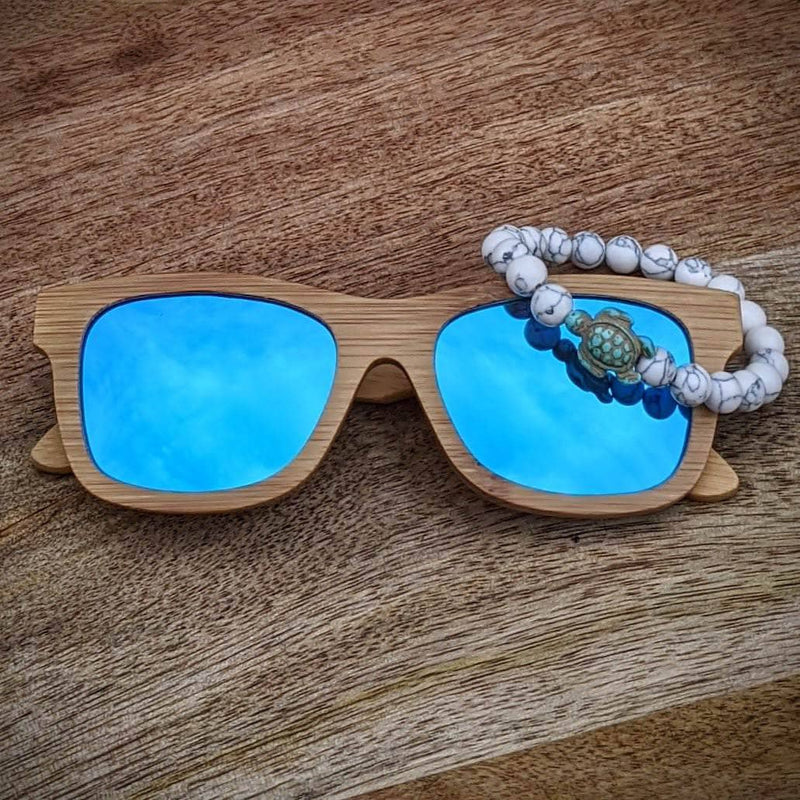 Blue mirror lens bamboo sunglasses paired with a white turquoise turtle bracelet, displayed on a wooden surface with a natural grain.
