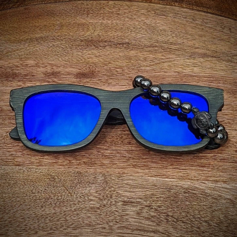 Blue mirror lens dark bamboo sunglasses paired with a polished hematite turtle bracelet, displayed on a round wooden surface.
