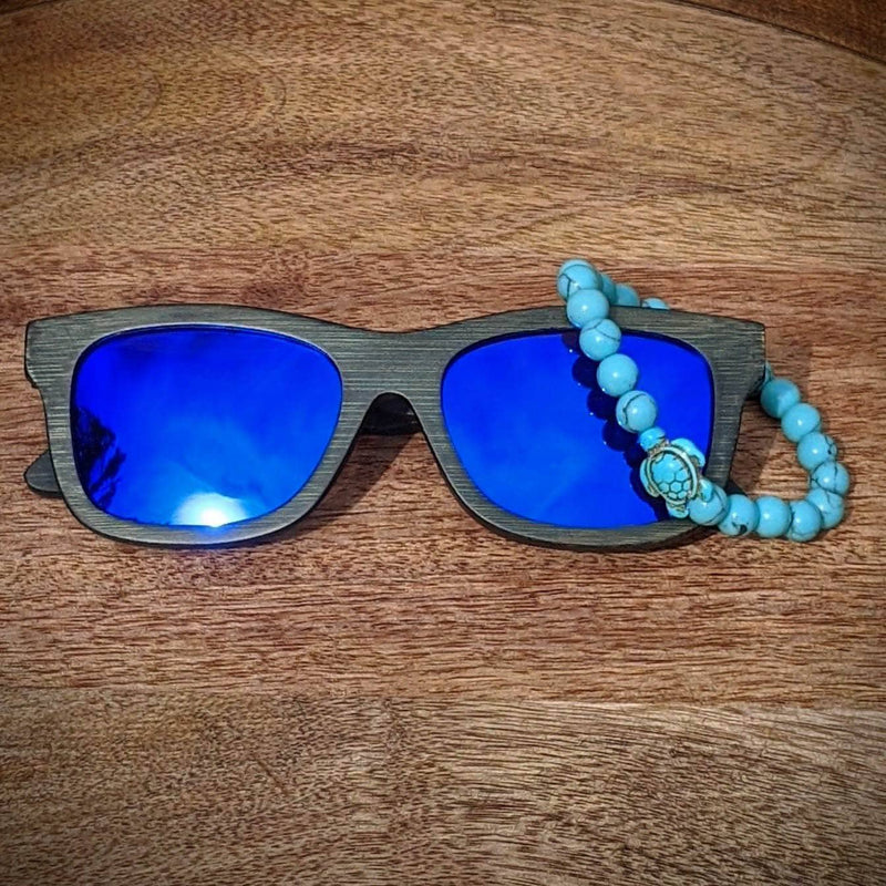 Blue mirror lens dark bamboo sunglasses paired with a turquoise gemstone turtle bracelet, displayed on a circular wooden tray.
