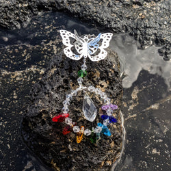 Butterfly crystal sun catcher with chakra beads resting on natural stone near water.