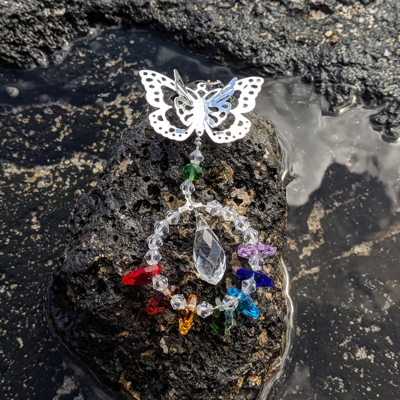 Butterfly crystal sun catcher with chakra beads resting on natural stone near water.