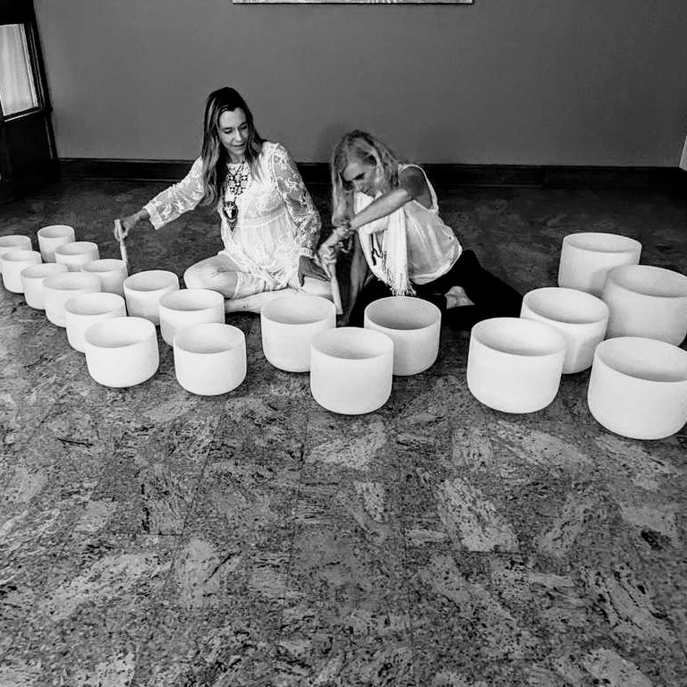 Two women playing a full chakra crystal bowl set on the floor during a healing session.