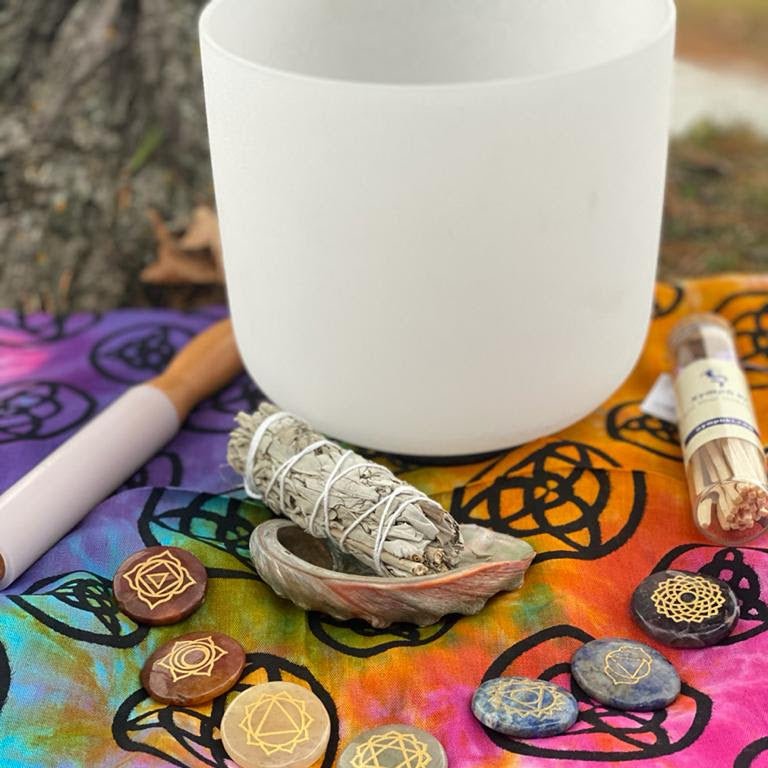 Singing bowl surrounded by chakra stones, sage stick, and abalone shell on patterned fabric.