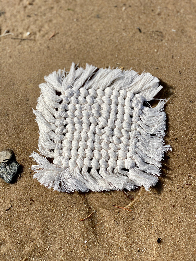 Set of four handcrafted macrame coasters with fringe detailing, displayed on a sandy beach.