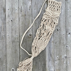 Handwoven macrame yoga mat bag in natural cord, hanging on a driftwood hook outdoors in sunlight.

