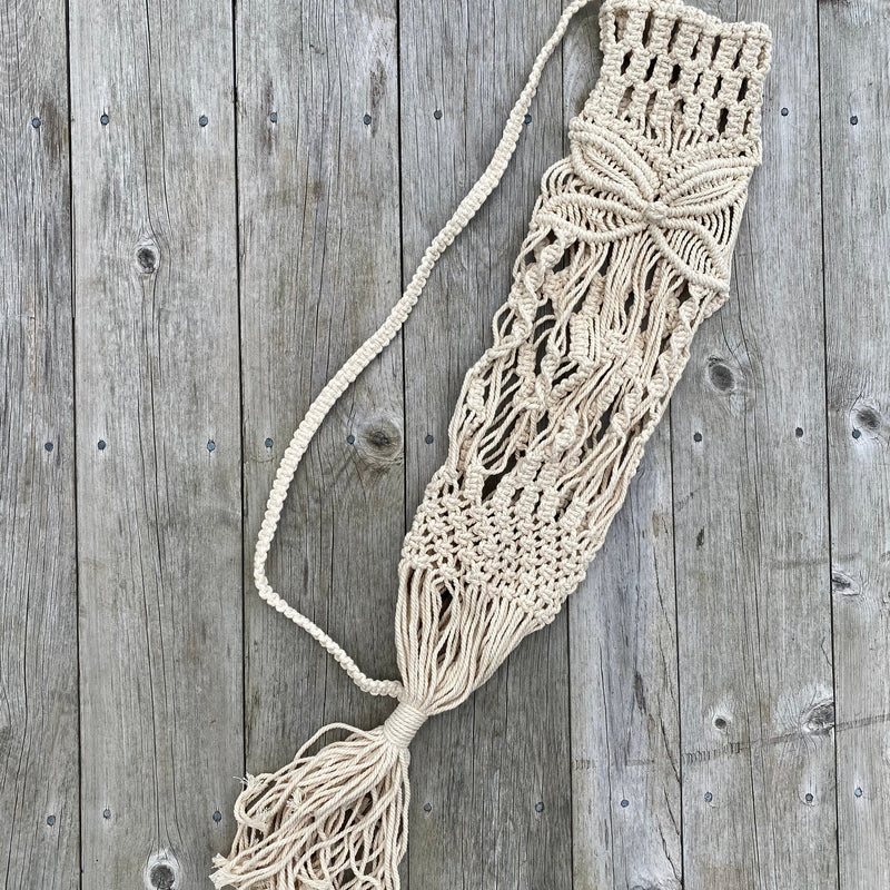 Handwoven macrame yoga mat bag in natural cord, hanging on a driftwood hook outdoors in sunlight.

