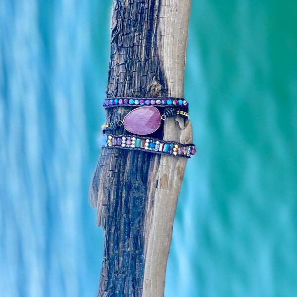 Magenta Wrap Bracelet with pink center stone and multi-color crystals, wrapped around driftwood against a teal water backdrop.