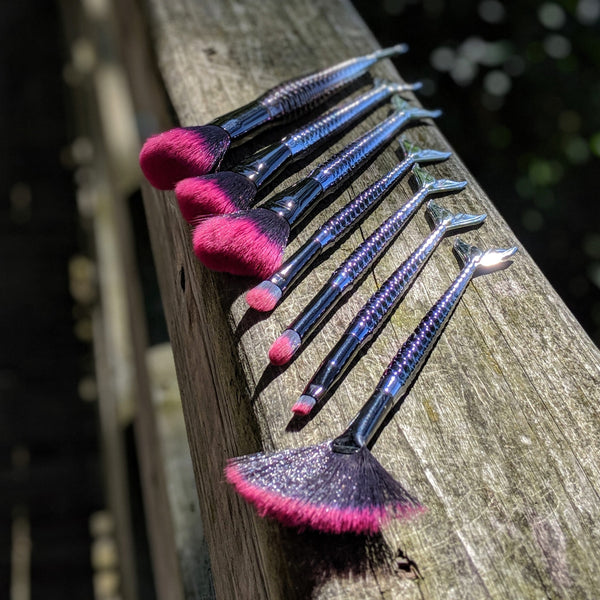 Set of mermaid makeup brushes with pink tips and shimmering handles, lined up on a rustic wooden ledge.