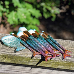 Blue-tipped mermaid makeup brushes on a wooden railing under bright sunlight.