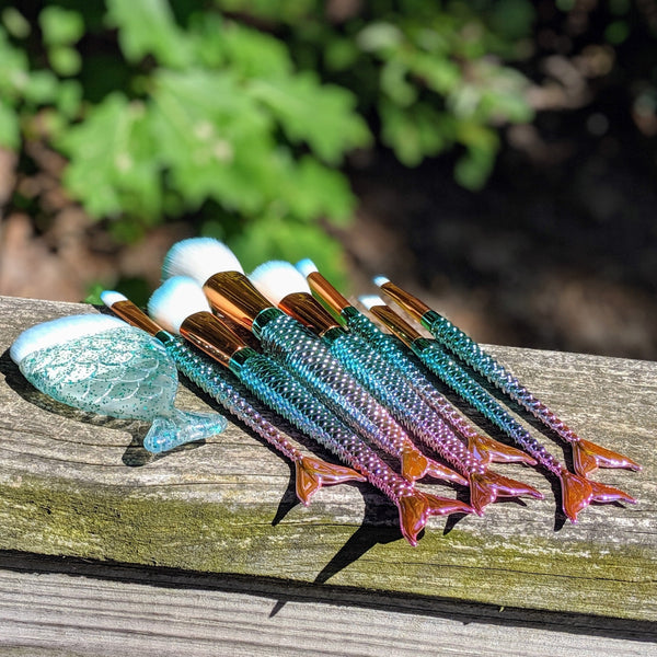 Blue-tipped mermaid makeup brushes on a wooden railing under bright sunlight.