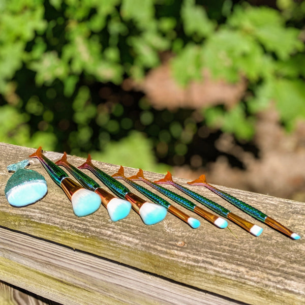 Blue-tipped mermaid makeup brushes fanned out on a wooden railing under bright sunlight.