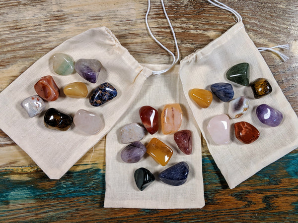 Assorted polished chakra stones spread around two cotton pouches, each with a rainbow of crystal colors.