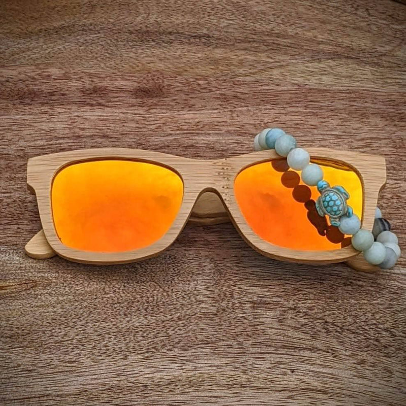 Orange mirror lens bamboo sunglasses paired with a druzy agate turtle bracelet, displayed on a warm wooden surface.

