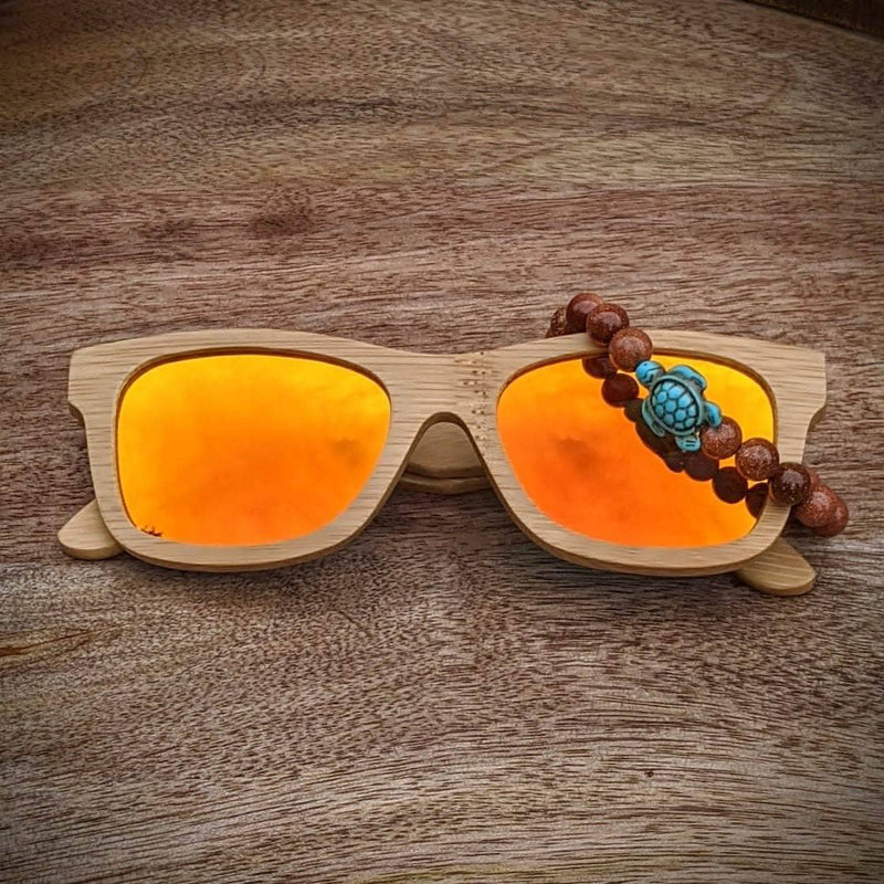 Orange mirror lens bamboo sunglasses paired with a gold sandstone turtle bracelet, displayed on a textured wooden surface.
