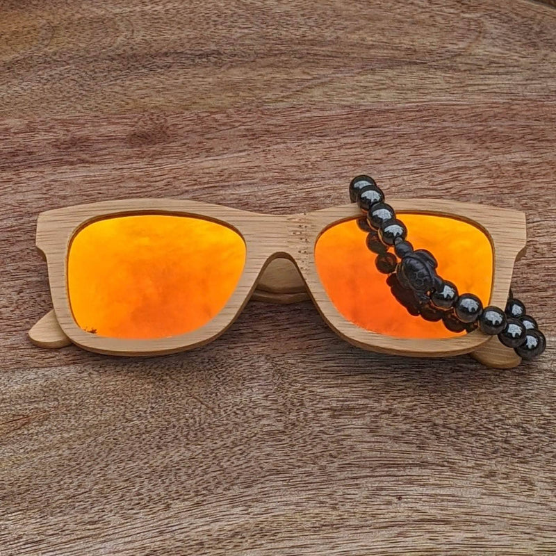 Orange mirror lens bamboo sunglasses paired with a polished hematite turtle bracelet, displayed on a smooth wooden background.
