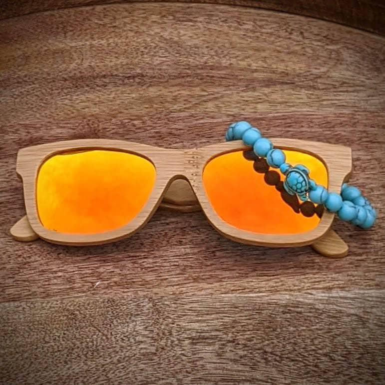Orange mirror lens bamboo sunglasses paired with a turquoise gemstone turtle bracelet, displayed on a smooth wooden surface.
