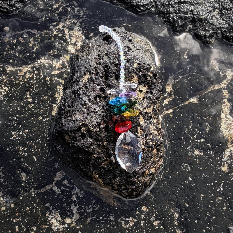 Oval-shaped chakra sun catcher with rainbow prism and chakra crystal beads displayed on natural stone background.