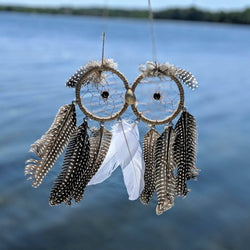 Owl-shaped dreamcatcher with spotted feathers and a white accent feather hanging over a calm lake.
