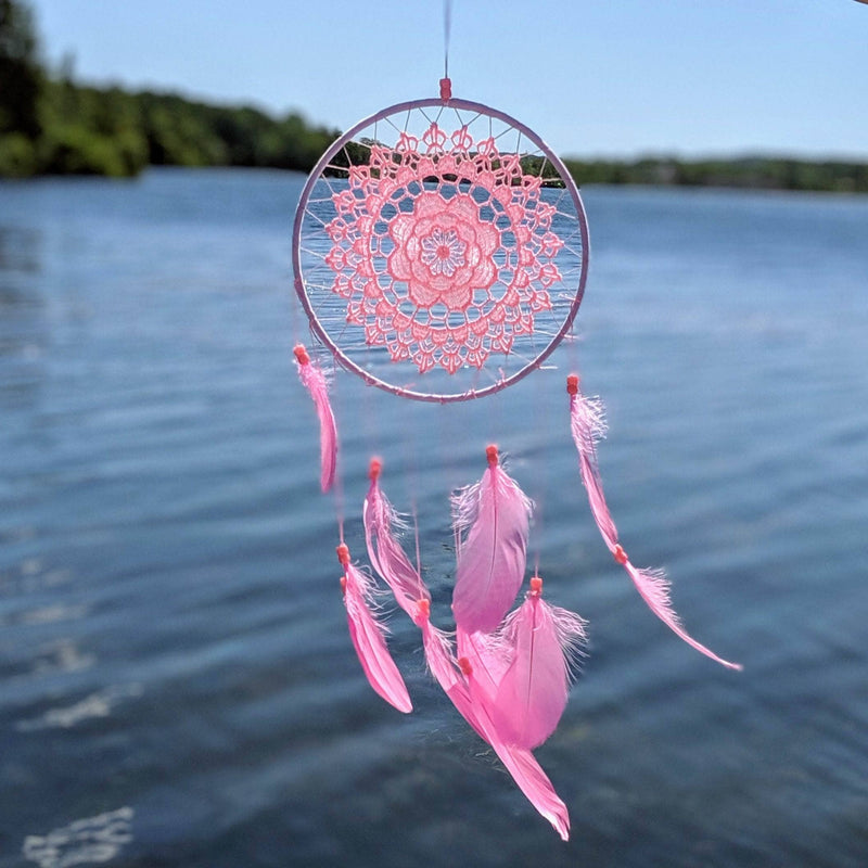 Pink lace dreamcatcher with feathers hanging above a serene lake on a clear sunny day.
