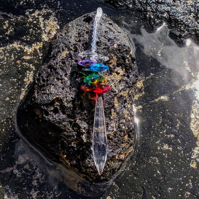 Pointed crystal sun catcher with chakra-colored beads placed on a stone near water.